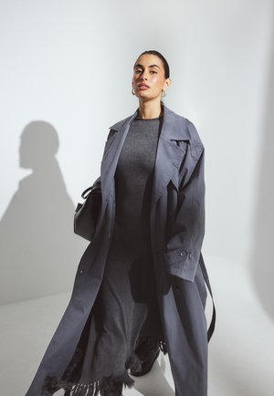 Naine kannab pikka halli trench coat'i tumeda halli kleidi üle, millel on frantsiääriga ääred, seistes ühetoonilise valge tausta ees.