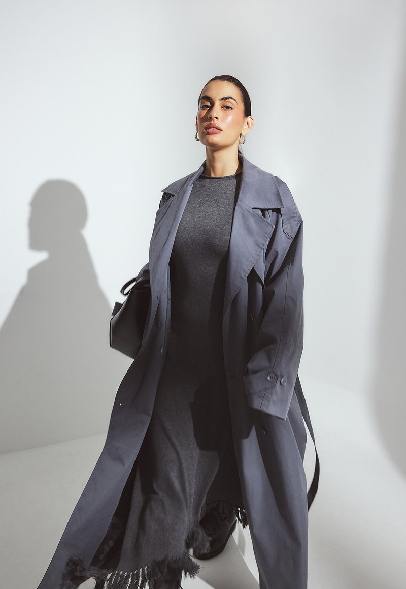 Naine kannab pikka halli trench coat'i tumeda halli kleidi üle, millel on frantsiääriga ääred, seistes ühetoonilise valge tausta ees.