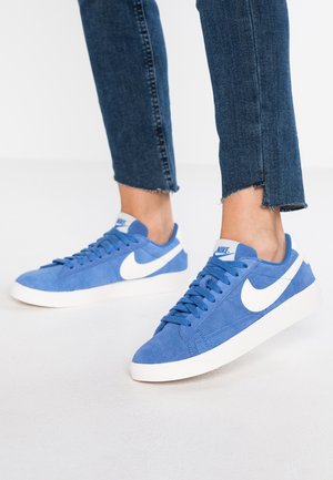 Niebieskie zamszowe tenisówki z białym logo Nike, o niskim profilu, płaskiej białej podeszwie i teksturowanych sznurowadłach. Dżinsy z postrzępionymi mankietami.