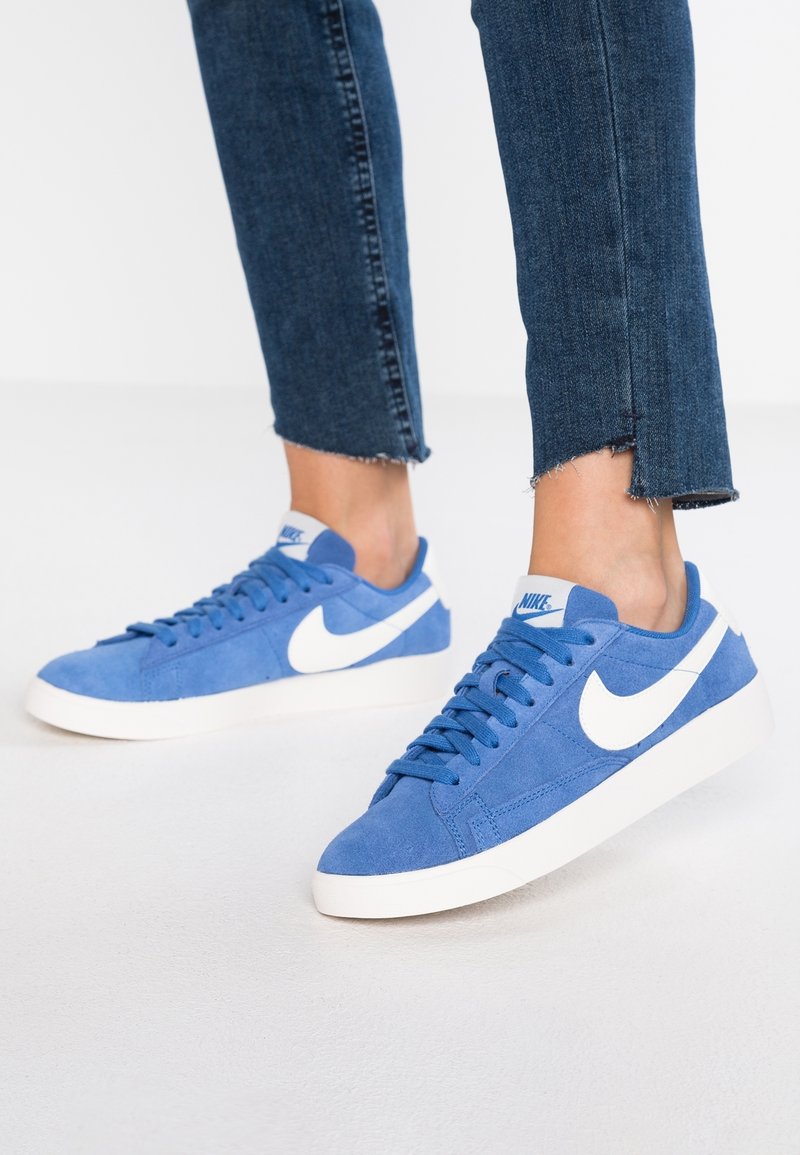 Baskets en daim bleu avec le logo Nike blanc en forme de virgule, présentant un profil bas, une semelle plate blanche et des lacets texturés. Jean en denim avec ourlets effilochés.