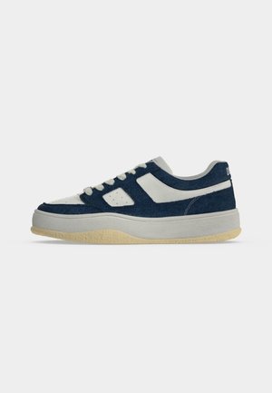 Zapatilla baja con paneles blancos y azul oscuro, cordones blancos y una gruesa suela beige, mostrada de perfil sobre un fondo liso.