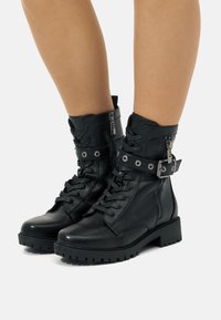 Botas de tobillo de cuero negro con punta redondeada, parte delantera con cordones, cremallera lateral, correa de hebilla ajustable y suela de goma gruesa.