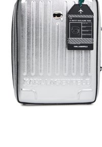 Srebrna walizka o twardej powłoce z pionowymi żłobieniami, wytłoczone logo Karla Lagerfelda, zawieszka w kształcie twarzy z okularami przeciwsłonecznymi oraz czarny tag z danymi podróży.