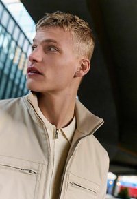 Beige colbert met ritssluiting en twee voorzakken. Daaronder is een lichtgekleurde, kortemouwen shirt zichtbaar.
