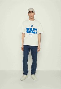 Balta medvilnė marškinėliai su mėlynu grafiniu tekstu, derinami su tamsiai mėlynomis džinsais ir baltais sportbačiais. Modelis dėvi kreminį kepuraitę. Paprasta fono.