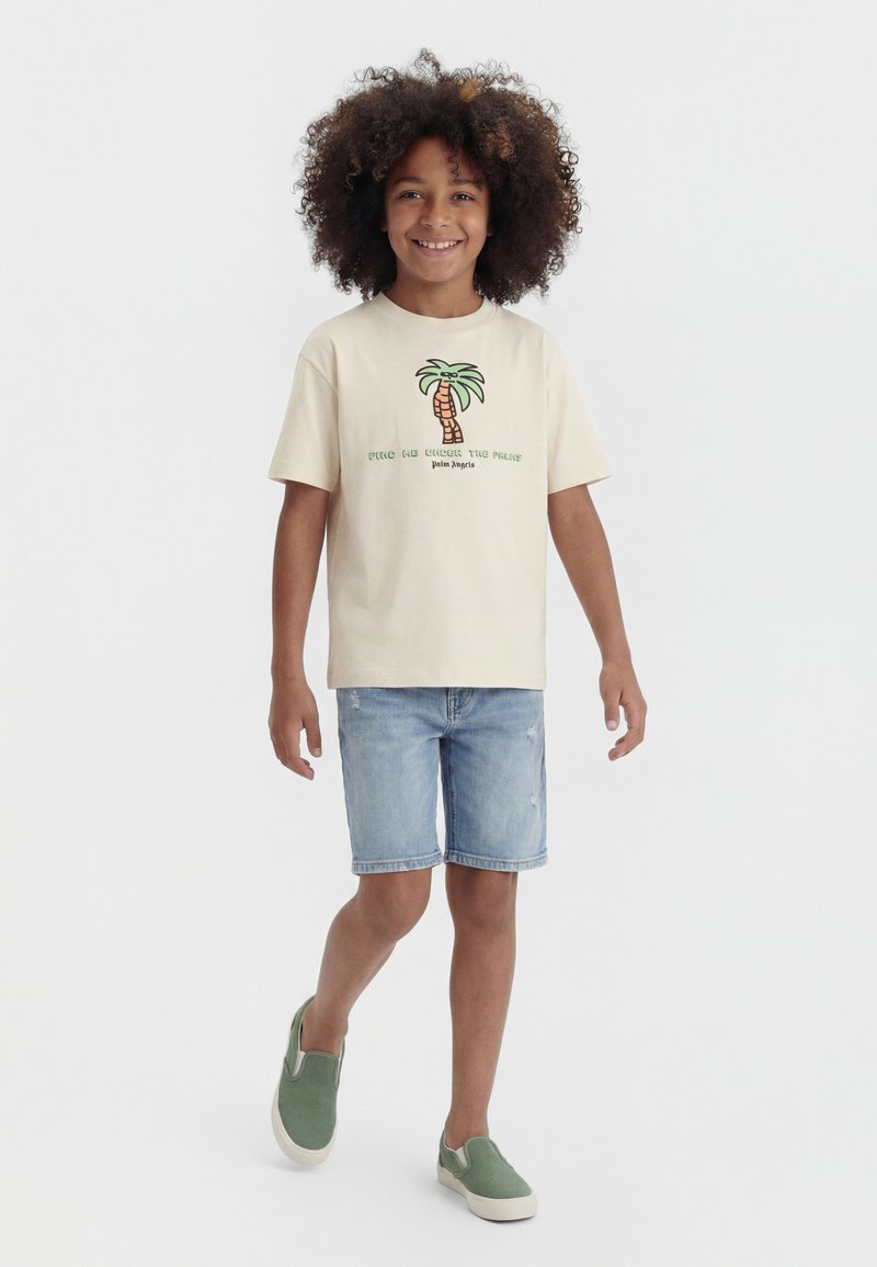 Child with curly hair walking, wearing a beige T-shirt with palm tree graphic, light denim shorts, and green slip-on shoes.