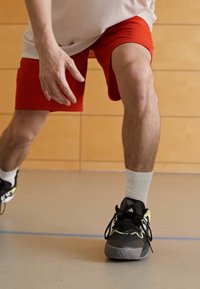 Svart joggesko med gule detaljer, iført hvite ankelsokker, røde shorts og en hvit mesh-genser, plassert på et glatt grått gulv.