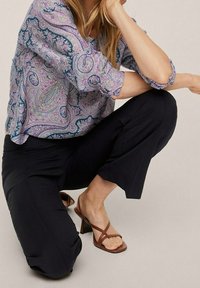 Blouse à motif cachemire violet avec manches retroussées, associée à un pantalon noir et des sandales à talons avec brides marron. Coupe décontractée et confortable.