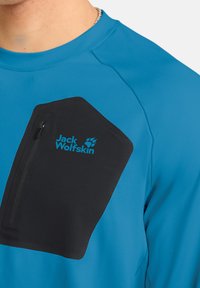 Blaues Technisches Shirt mit einer schwarzen Reißverschlusstasche auf der Brust und dem blauen "Jack Wolfskin"-Logo. Glatte Stoffqualität mit taillierter Passform.