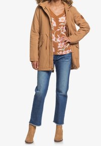 Parka beige con cappuccio foderato di pelliccia, vita stretta e due tasche frontali; abbinata a un top a fantasia floreale, jeans blu e stivali alla caviglia beige.
