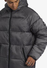 Grå pufferjacka med huva, med quiltad design, framdragkedja och sidofickor. Tillverkad av glänsande, lätt material.