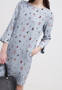 Grafikowa sukienka z jasnoniebieskiej tkaniny z okrągłymi i prostokątnymi wzorami biżuteryjnymi w różnych kolorach. Posiada rękawy 3/4 oraz boczne kieszenie.