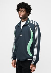 Μπλε σκούρο μπουφάν με φερμουάρ και πράσινες λεπτομέρειες, λευκές ρίγες στα μανίκια και κεντημένο λογότυπο. Διαθέτει ψηλό γιακά και ελαστικά μανσέτα.