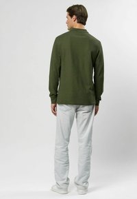 Olivovo zelená košeľa s dlhým rukávom, hladká textura, voľný strih, skombinovaná so svetlo šedými džínami a bielymi topánkami, z pohľadu zozadu.