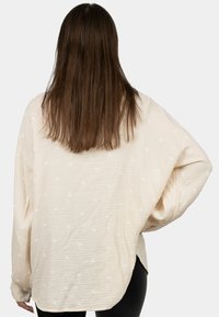 Camicia oversize beige chiaro realizzata in tessuto testurizzato con pois bianchi, caratterizzata da maniche ampie e orlo curvo. Vista posteriore mostrata.