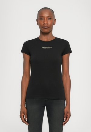 Vrouw met kort haar die een nauwsluitend zwart Armani Exchange t-shirt en donkere spijkerbroeken draagt, staande tegen een effen lichte achtergrond.
