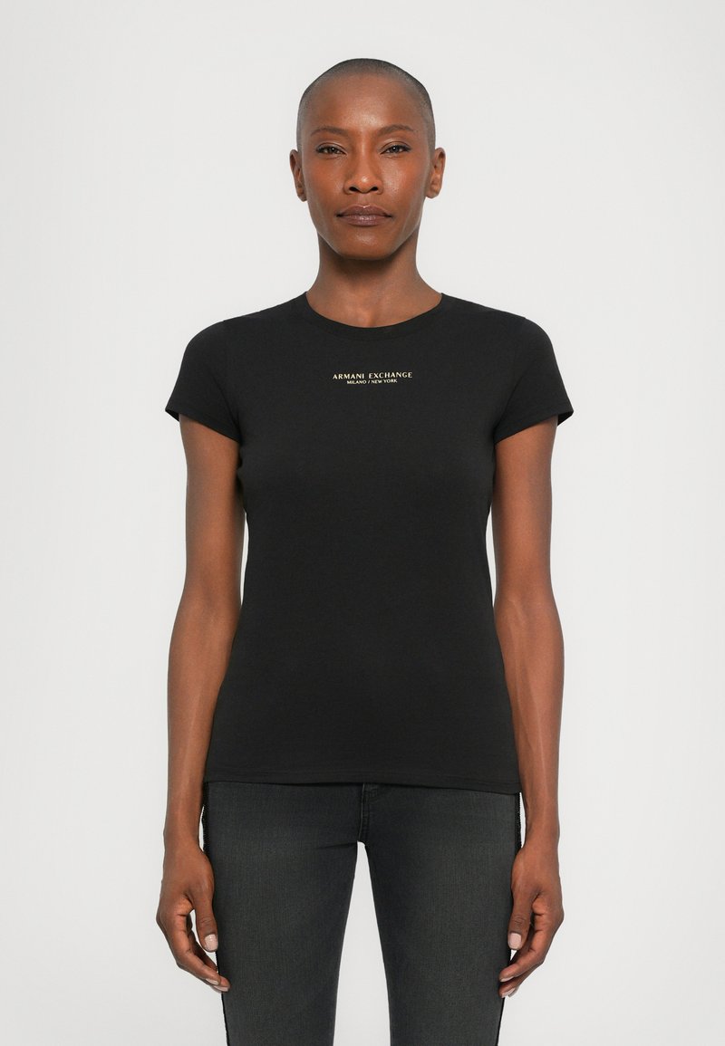 Vrouw met kort haar die een nauwsluitend zwart Armani Exchange t-shirt en donkere spijkerbroeken draagt, staande tegen een effen lichte achtergrond.