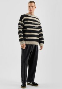 Sweter w paski w kolorach czarnym i beżowym z prążkowanym, okrągłym dekoltem, zestawiony z luźnymi czarnymi spodniami i czarnymi trampkami z białymi akcentami.