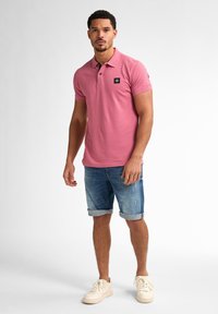 Polo en coton rose avec un patch logo noir, associé à un short en denim bleu et des baskets blanches. Coupe décontractée, manches courtes, shorts retroussés.