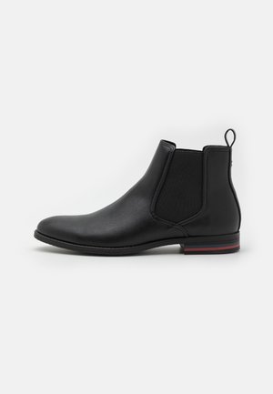 Bottine Chelsea noire en cuir avec panneaux latéraux élastiques, languette au talon et semelle plate avec un détail subtil de rayure rouge.