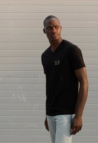 Μαύρο t-shirt με V λαιμόκοψη και στενή εφαρμογή, με μικρό, χρυσό τετράγωνο λογότυπο στο αριστερό στήθος. Φορεμένο με ανοιχτό μπλε τζιν.