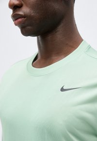 Κοντινό πλάνο ενός ατόμου που φοράει ένα ανοιχτό πράσινο μπλουζάκι Nike με μαύρο λογότυπο Nike swoosh στην αριστερή πλευρά του στήθους.