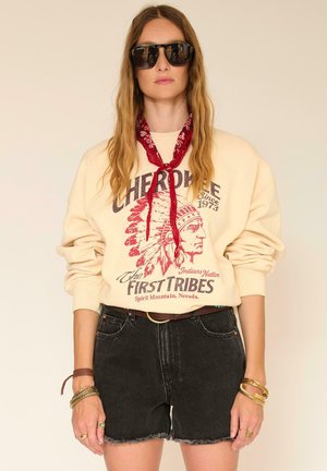 Femme portant un sweat-shirt crème oversize avec un imprimé amérindien, un short en jean noir, un bandana rouge, de grandes lunettes de soleil noires et des bracelets en or.