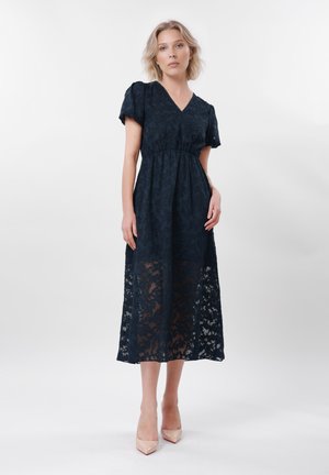 Mujer de pie con un vestido midi negro de escote en V, mangas cortas y superposición de encaje floral transparente, combinado con tacones beige de punta afilada.