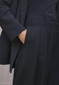Abito scuro su misura con giacca strutturata e pantaloni plissettati. Tessuto morbido, linee nette e design minimalista con tasche profonde.