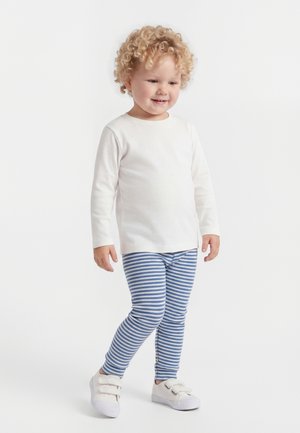 Väikelaps lokkis juustega, riietatud valgesse pika varrukaga särki, sinise ja valge triibulisse retuusidesse ning valgetesse velkkingadesse, seisab ja naeratab.