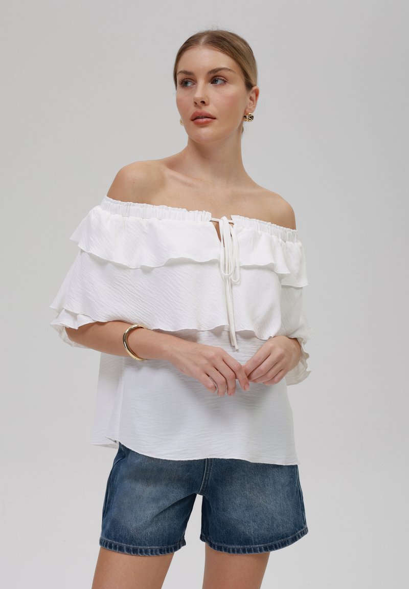 Witte off-the-shoulder top met geruite lagen, trekkoord bij de hals, gecombineerd met blauwe denim shorts. Zachte, gestructureerde stof met een lichte val.