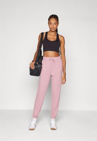Ružové tepláky s elastickými manžetami a sťahovacím pásom, v kombinácii s čiernym cropped topom. Modelka nesie čiernu športovú tašku.