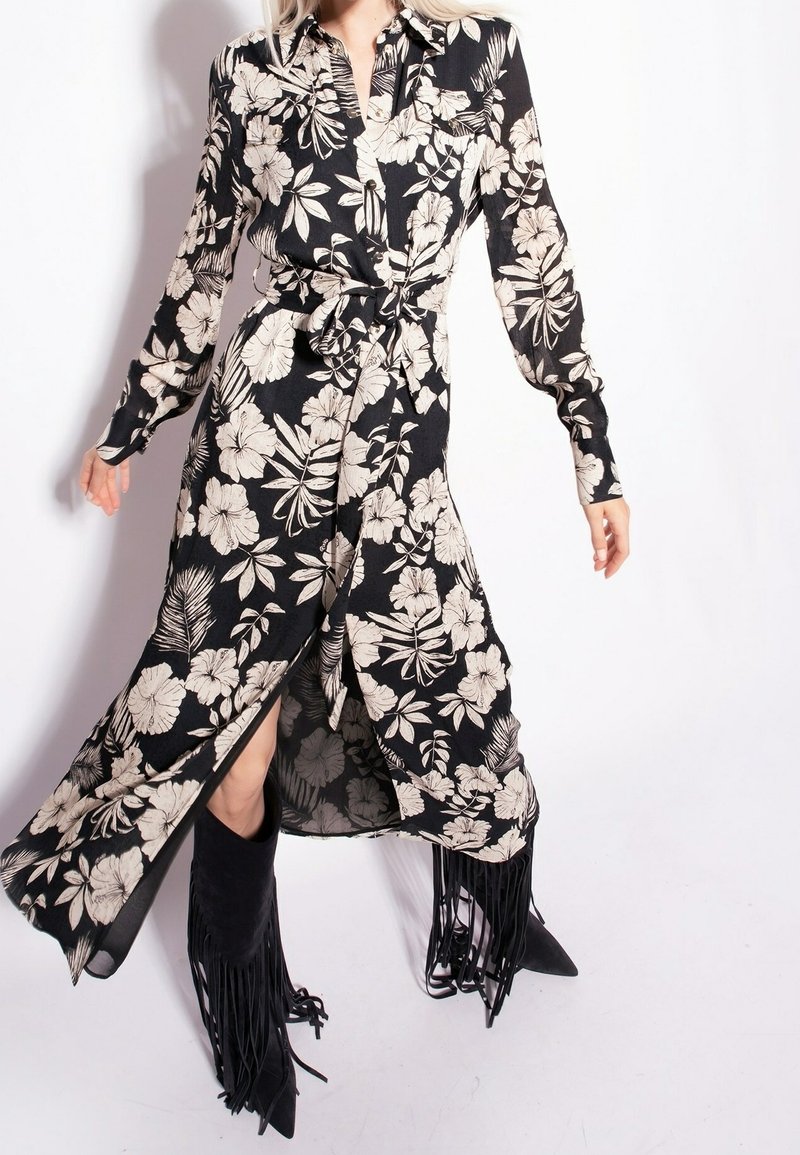 Kobieta ubrana w czarną kwiecistą sukienkę midi z przewiązką w talii oraz czarne, frędzlowane botki na szpilce, pozująca na tle białym.