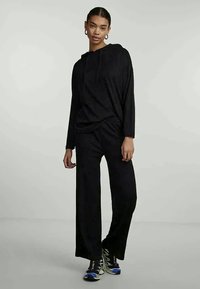 Μαύρη ριγωτή μπλούζα με κουκούλα και μακριά μανίκια, συνδυασμένη με άνετα μαύρα παντελόνια, υφή υφάσματος και χοντρές αθλητικές γνωστές με μπλε λεπτομέρειες.