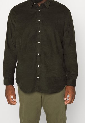 Mężczyzna ubrany w ciemnozieloną koszulę na długi rękaw z guzikami oraz oliwkowozielone spodnie, stojący na tle jednolitego, jasnego tła.