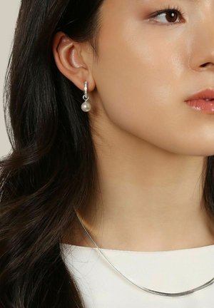 Silvriga hängande örhängen med en rund pärla och kristall-accenter. Bäraren har vågigt mörkt hår och ett enkelt silvrigt halsband.