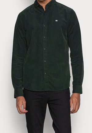 Mann trägt ein dunkelgrünes, langärmeliges Hemd mit Knöpfen und einem kleinen weißen bestickten Logo auf der linken Brust und schwarze Hose.