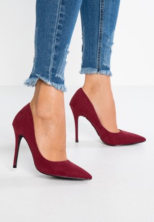 Escarpins à talons hauts en daim bordeaux, à bout pointu et silhouette élégante, associés à un jean en denim déchiré avec des ourlets effilochés.