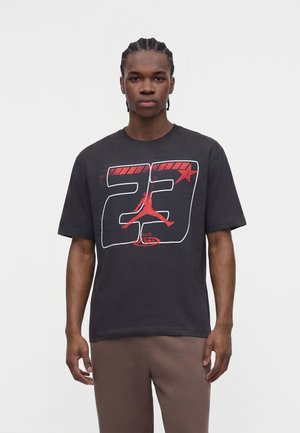 Mladić s pletenicama nosi crnu majicu s crvenom i bijelom grafikom "23" s jumpman siluetom i zvijezdom.