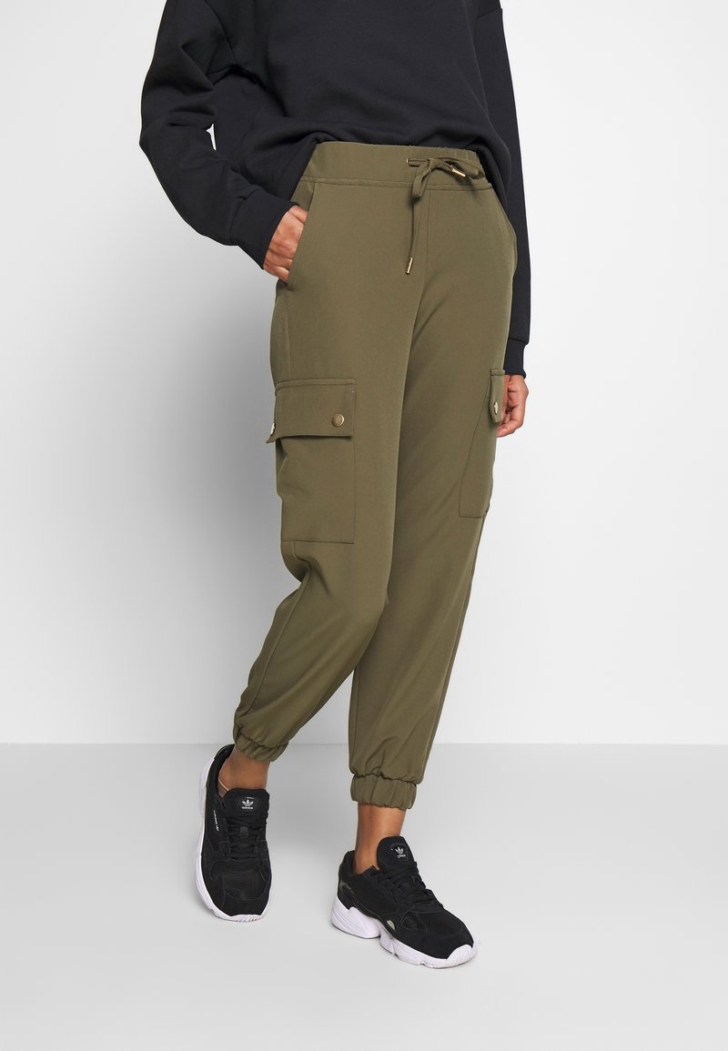 Personne portant un pantalon de jogging vert olive avec taille à cordon et grandes poches latérales boutonnées, associé à des baskets noires et un haut noir à manches longues.