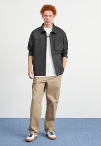 Sort struktureret skjorte med knapper og en brystlomme, båret over en hvid t-shirt, parret med beige cargo-bukser og hvide sneakers med marineblå snørebånd.
