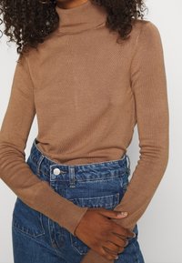 Brązowy golf wykonany z miękkiego, prążkowanego materiału. Długie rękawy, dopasowany fason. Połączony z wysokim stanem niebieskich dżinsów.