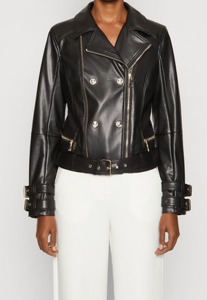 Kobieta w czarnej, dwurzędowej, skórzanej kurtce motocyklowej z srebrnymi zamkami błyskawicznymi, zapięciami na mankietach i paskiem, w połączeniu z białymi spodniami.