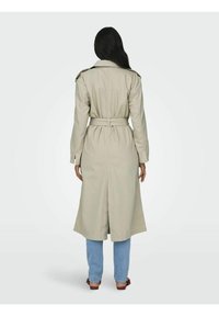 Beige trenchcoat met lange mouwen, ceintuurlijn, en een rugventilatie. Gecombineerd met lichtblauwe jeans en bruine sandalen. Gladde textuur.