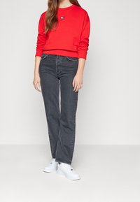 Rode sweatshirt met logo patch, gecombineerd met donkergrijze high-waisted jeans en witte sneakers. Gladde textuur, relaxte fit en eenvoudig ontwerp.