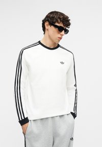 Jeune homme portant un t-shirt blanc Adidas à manches longues avec des bandes noires et un pantalon de survêtement gris Adidas, mains dans les poches, regardant vers le bas et portant des lunettes de soleil noires.