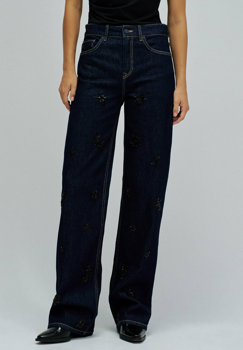 Jean en denim foncé à jambes larges avec des motifs de fleurs embellis. Présente des coutures contrastantes et une taille haute. Fini avec une fermeture à bouton classique.