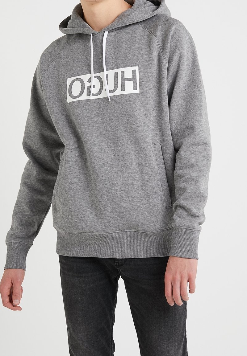 Personne portant un sweat à capuche gris avec des cordons blancs et un logo blanc en bloc avec "HUGO" imprimé à l'envers sur la poitrine, associé à un jean noir.