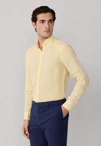Homme portant une chemise à manches longues jaune clair boutonnée, rentrée dans un pantalon bleu marine, debout avec une main dans la poche.