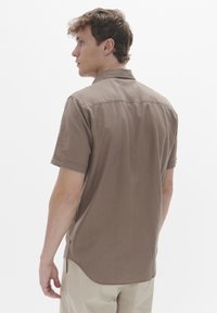 Cămașă cu mânecă scurtă, de culoare maro, fabricată dintr-un material moale, având un guler clasic, o cusătură pe spate și un tiv curbat, cu detalii minime de coasere.